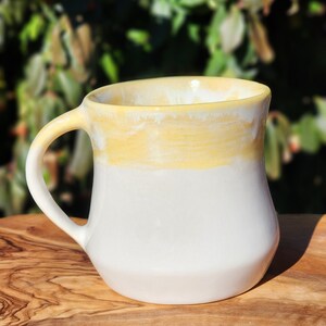 May include: A white ceramic mug with a yellow glaze on the top half. The mug has a rounded handle and a slightly flared lip.