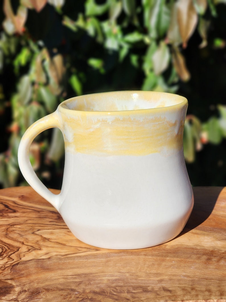 May include: A white ceramic mug with a yellow glaze on the top half. The mug has a rounded handle and a slightly flared lip.