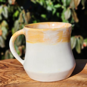 May include: A white ceramic mug with a yellow glaze on the top half. The mug has a rounded body and a handle.