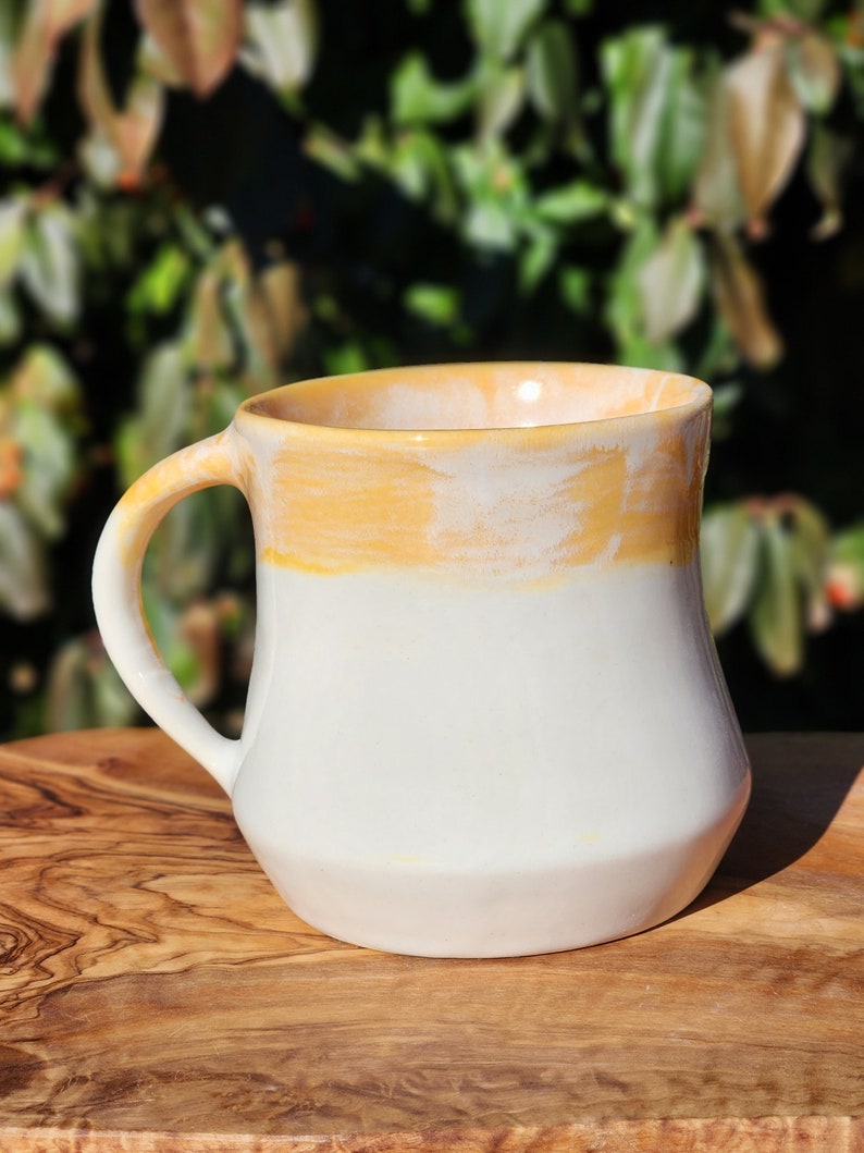 May include: A white ceramic mug with a yellow glaze on the top half. The mug has a rounded body and a handle.