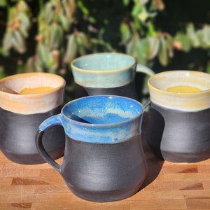 Op de afbeelding: Vier handgemaakte keramische mokken met een zwarte basis en gekleurde randen. De mokken hebben een matte zwarte buitenkant en een glanzende binnenkant met kleuren als blauw, geel en teal. De mokken staan op een houten snijplank.