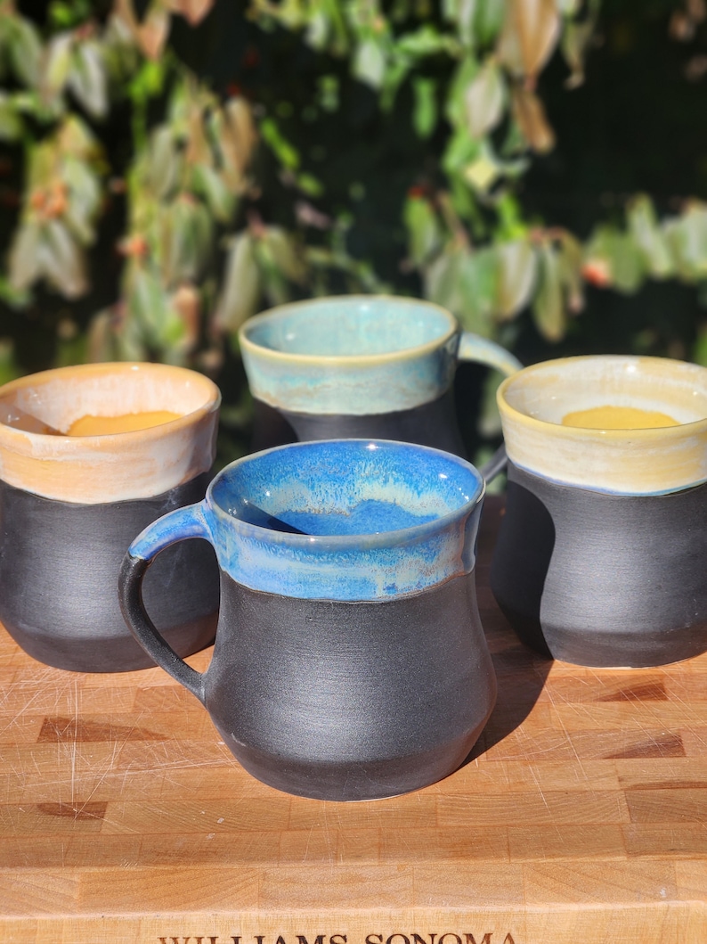 Op de afbeelding: Vier handgemaakte keramische mokken met een zwarte basis en gekleurde randen. De mokken hebben een matte zwarte buitenkant en een glanzende binnenkant met kleuren als blauw, geel en teal. De mokken staan op een houten snijplank.
