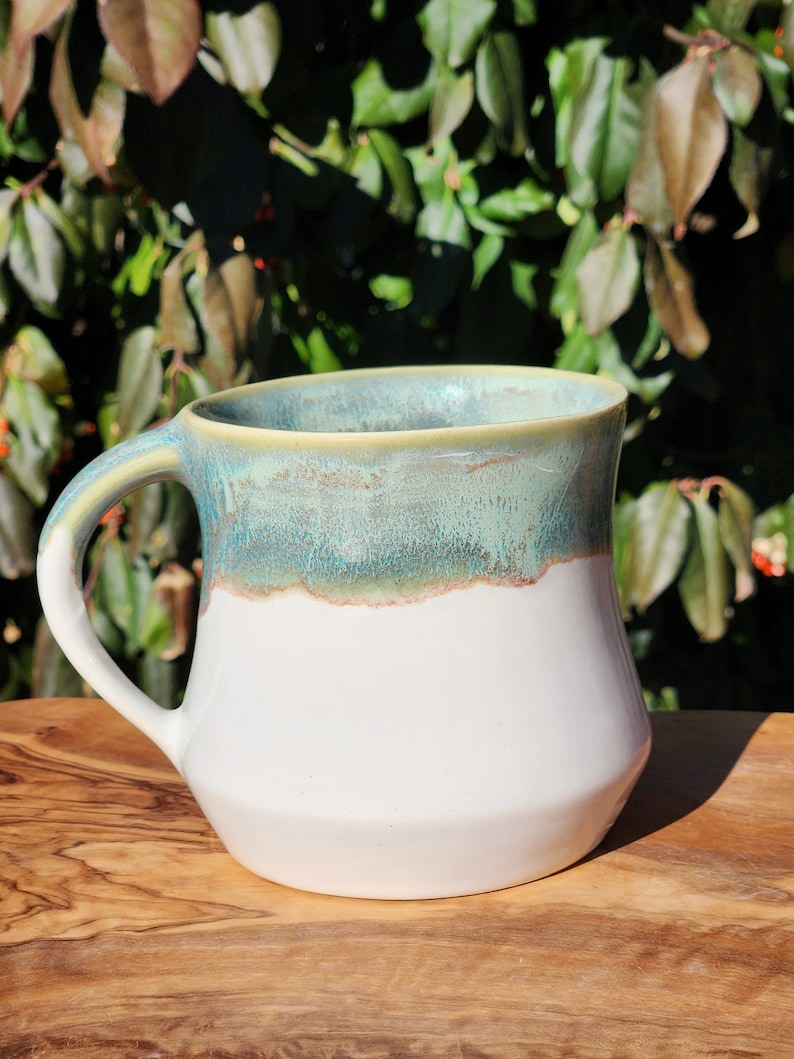 May include: A white ceramic mug with a teal and green glaze. The mug has a wide base and a rounded handle.