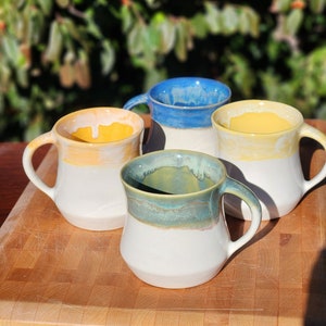 May include: Four ceramic mugs with a white base and a colored rim. The mugs are yellow, blue, and green. The mugs are sitting on a wooden cutting board.
