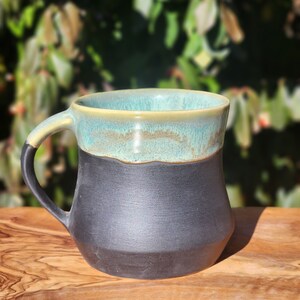 Op de afbeelding: Een handgemaakte keramische mok met een zwarte basis en een turquoise rand. De handgreep is zwart en de binnenkant is lichtbeige. De mok staat op een houten oppervlak met een vage groene achtergrond.