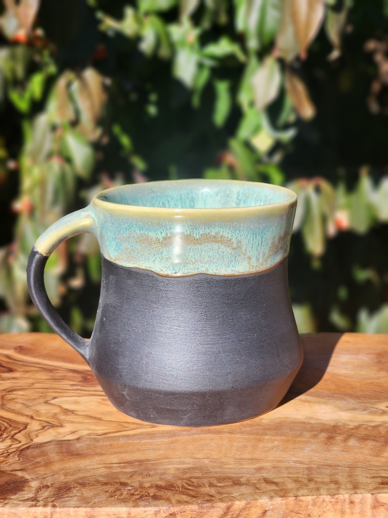Op de afbeelding: Een handgemaakte keramische mok met een zwarte basis en een turquoise rand. De handgreep is zwart en de binnenkant is lichtbeige. De mok staat op een houten oppervlak met een vage groene achtergrond.