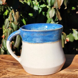 May include: A white ceramic mug with a blue glaze on the top half. The mug has a rounded body and a handle.