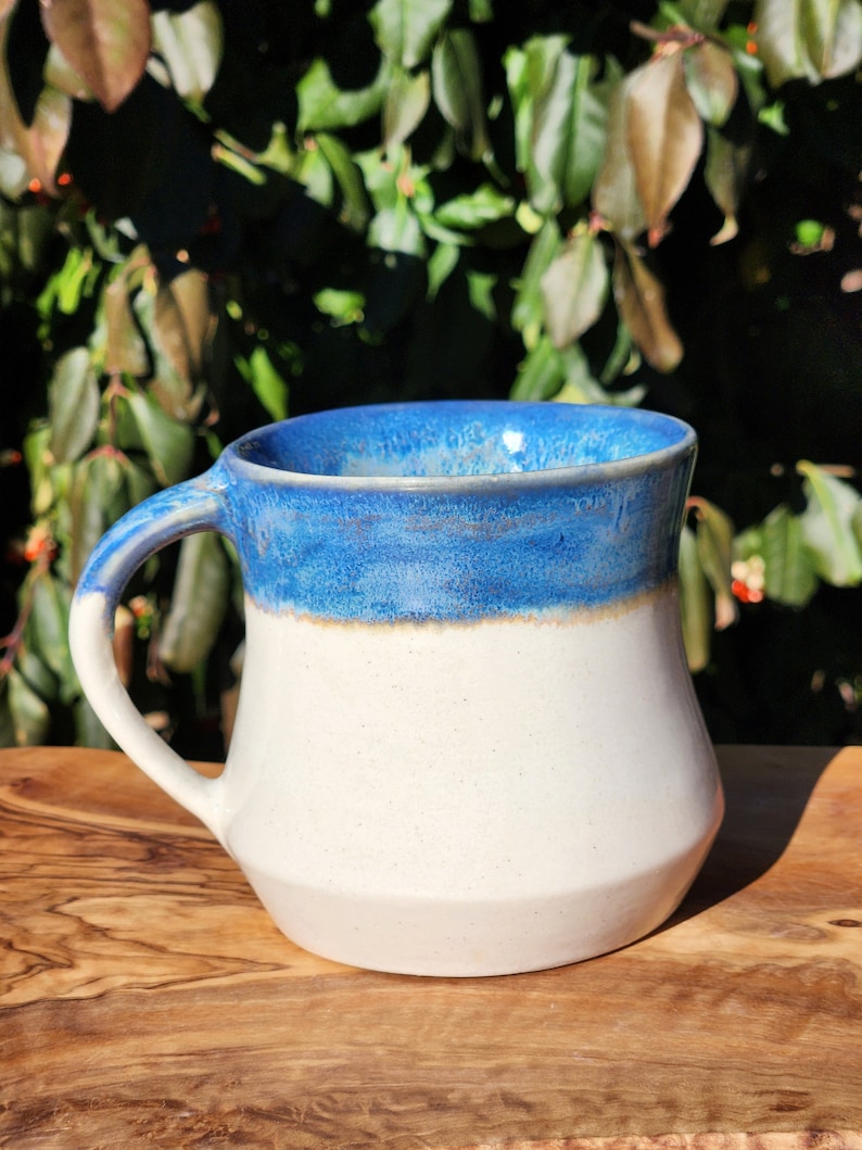 May include: A white ceramic mug with a blue glaze on the top half. The mug has a rounded body and a handle.