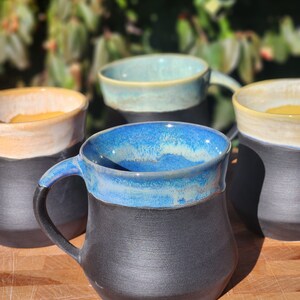 Op de afbeelding: Een verzameling handgemaakte keramische mokken. Elke mok heeft een donkere, getextureerde basis en een contrasterende, geglazuurde rand in tinten blauw, blauwgroen en cr&egrave;me. De mokken hebben een stevig handvat en een rustieke, ambachtelijke uitstraling.