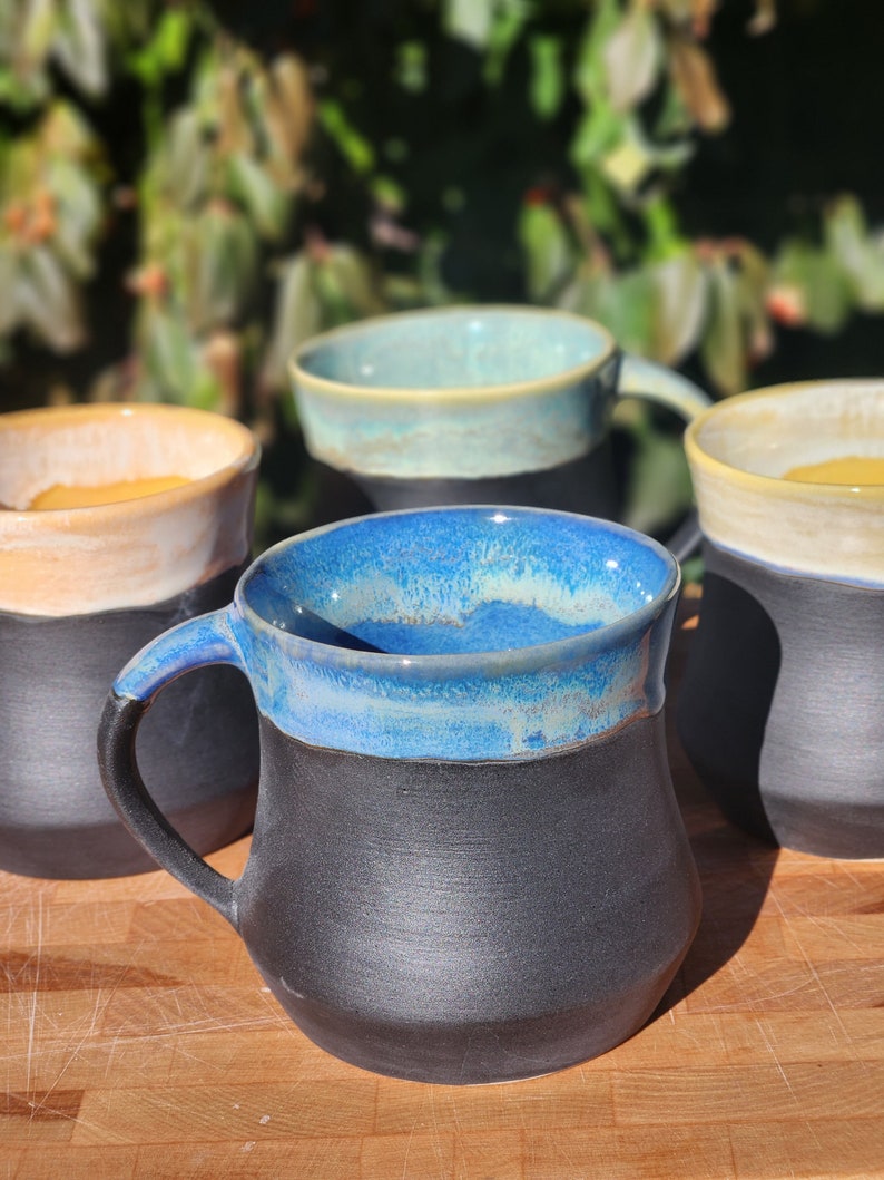 Op de afbeelding: Een verzameling handgemaakte keramische mokken. Elke mok heeft een donkere, getextureerde basis en een contrasterende, geglazuurde rand in tinten blauw, blauwgroen en cr&egrave;me. De mokken hebben een stevig handvat en een rustieke, ambachtelijke uitstraling.