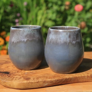May include: Two ceramic mugs with a dark blue and gray glaze. The mugs are sitting on a wooden cutting board.