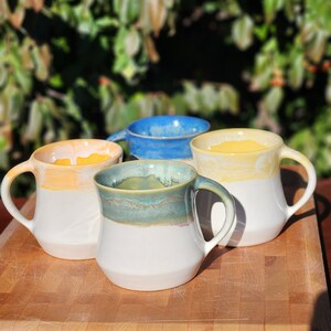 May include: Four ceramic mugs with different colored glazes. The mugs are white with a yellow, blue, and green glaze on the top. The mugs are sitting on a wooden cutting board.