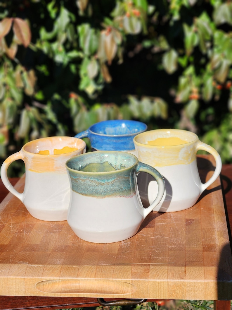 May include: Four ceramic mugs with different colored glazes. The mugs are white with a yellow, blue, and green glaze on the top. The mugs are sitting on a wooden cutting board.