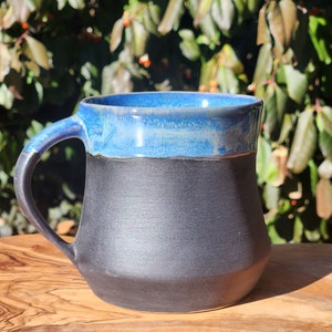 Op de afbeelding: Een handgemaakte keramische mok met een zwarte basis en een blauw geglazuurde rand en binnenkant. De mok heeft een gebogen handvat en staat op een houten oppervlak. De mok is ontworpen voor warme dranken.