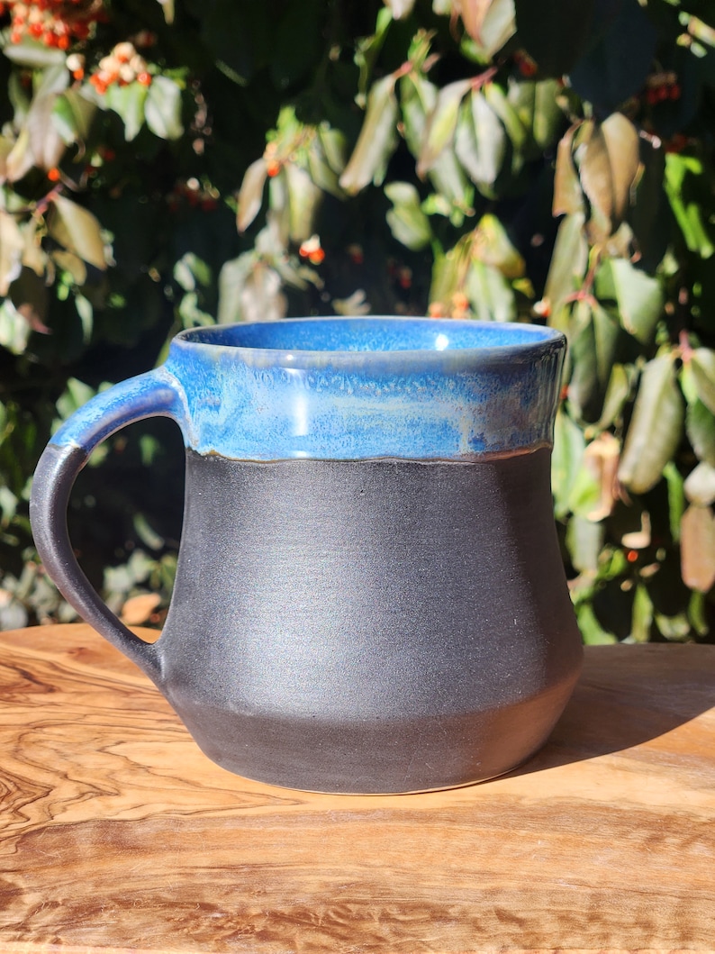 Op de afbeelding: Een handgemaakte keramische mok met een zwarte basis en een blauw geglazuurde rand en binnenkant. De mok heeft een gebogen handvat en staat op een houten oppervlak. De mok is ontworpen voor warme dranken.