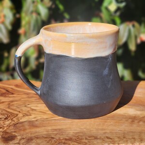 Op de afbeelding: Een handgemaakte keramische mok met een zwarte basis, een bruin/gele bovenkant en een witte rand. De mok heeft een gebogen handvat en staat op een houten oppervlak. De mok is ontworpen voor warme dranken.