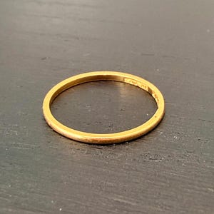 Puede incluir: Un anillo fino de color dorado. El anillo es circular y parece ser de metal. El anillo está sobre un fondo oscuro. El anillo es una banda simple, adecuada para el uso diario o para apilar con otros anillos.