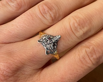May include: A gold ring featuring a detailed wolf head design. The wolf's head is encrusted with small, sparkling stones and has a red gemstone eye. The ring's band has a textured, twisted pattern.