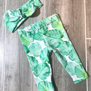 May include: A matching set of baby clothing featuring a tropical leaf pattern. The set includes a pair of leggings and a knotted headband. The leggings and headband are white with a watercolor design of large green leaves.