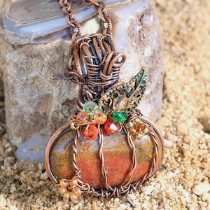 May include: A copper wire-wrapped pendant shaped like a pumpkin, featuring a central multicolored agate stone, accented with small green, orange, and red beads, and a decorative metal leaf, suspended from a matching copper chain.