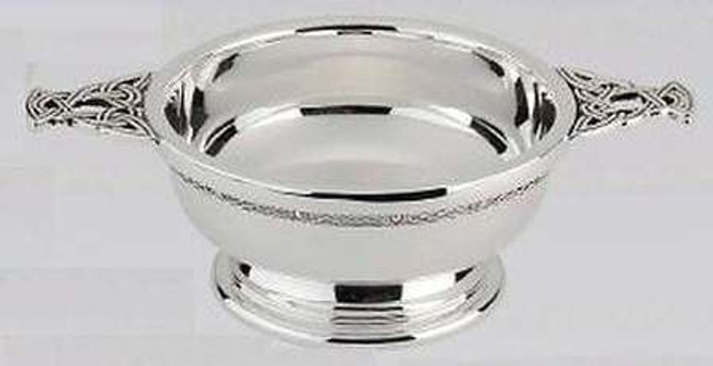 May include: A polished silver bowl with two decorative handles. The bowl has a patterned band around the rim and rests on a tiered base. The handles are adorned with Celtic knot designs.