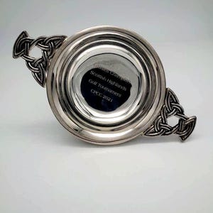 May include: A silver trophy with Celtic knot handles. The trophy has a circular bowl with the text "Classics Champion Scottish Highlands Golf Tournament CPCC 2021" engraved on the inside.