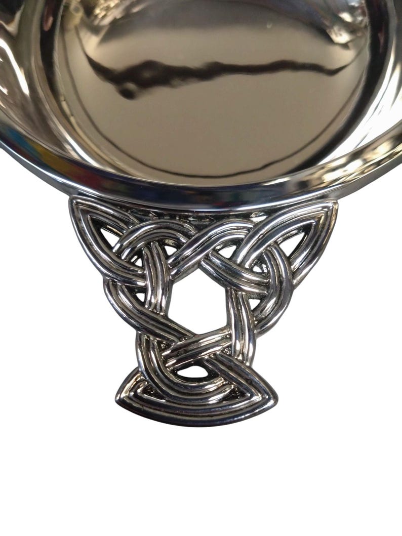 May include: A silver serving bowl with a Celtic knot handle. The bowl has a reflective surface, and the handle features an intricate, interwoven pattern. The bowl is likely made of metal. The bowl's diameter is approximately 20 cm.