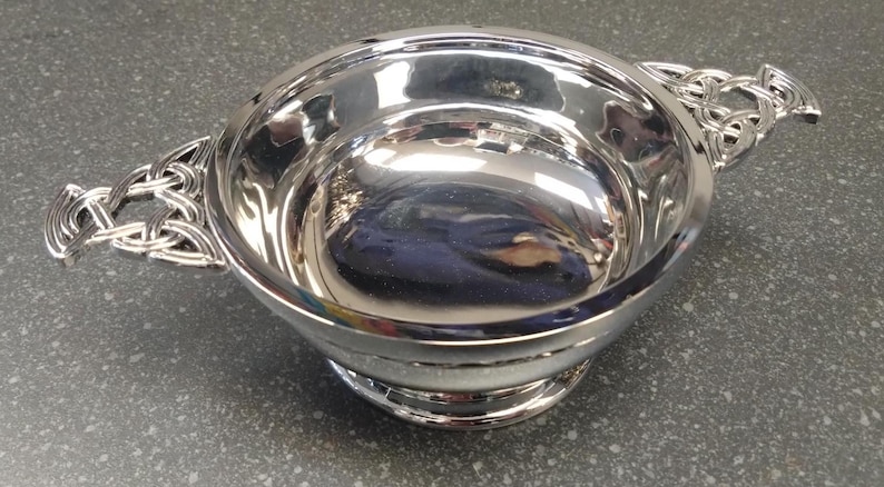 May include: A polished silver bowl with Celtic knot handles. The bowl is round with a small base and reflects the surroundings. The handles are intricately designed with a Celtic knot pattern.
