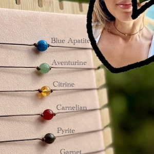 May include: A display of gemstone necklaces, each with a different colored bead. The gemstones include blue apatite, aventurine, citrine, carnelian, pyrite, garnet, and moonstone. The names of the gemstones are printed next to each necklace.