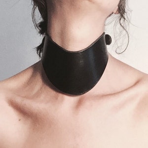 May include: A black leather choker collar with a wide, curved design. The collar fastens with a small, round buckle on the side. The choker sits around the neck, showcasing a minimalist and bold aesthetic.