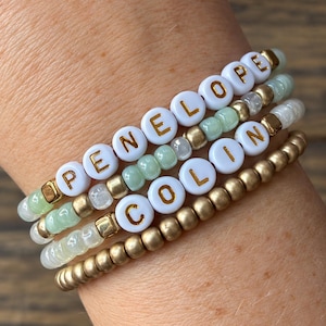 May include: A stack of four beaded bracelets featuring white round beads with gold lettering spelling out the names PENELOPE and COLIN, interspersed with light green, translucent, and metallic gold spherical beads on an elastic cord.