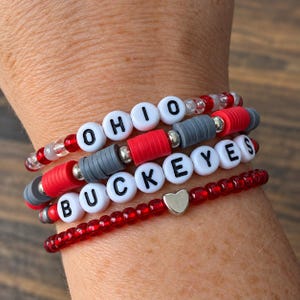 Könnte beinhalten: Ein Stapel aus vier Perlenarmbändern in den Farben Rot, Grau und Weiß. Ein Armband zeigt das Wort OHIO in schwarzen Buchstaben auf weißen Perlen, ein weiteres BUCKEYES und eines enthält einen kleinen silbernen herzförmigen Anhänger.