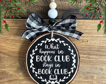May include: A wooden Christmas ornament with the text "What happens in BOOK CLUB stays in BOOK CLUB." The ornament is round, with a black painted surface and white lettering. It has a black and white checkered bow and is decorated with wooden beads.