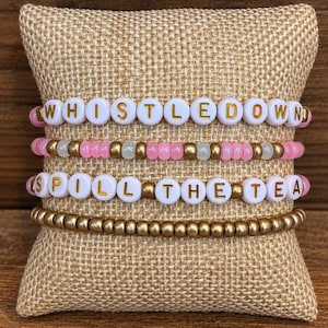 May include: A set of four beaded bracelets featuring white round beads with gold lettering that spells out WHISTLE DOWN and SPILL THE TEA, interspersed with metallic gold and translucent pink beads arranged in a stacked design on a textured pillow.