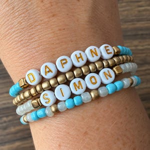 May include: A stack of four bracelets featuring white round beads with gold lettering spelling DAPHNE and SIMON, interspersed with metallic gold-tone spherical beads and translucent light blue and clear glass seed beads on elastic cord.
