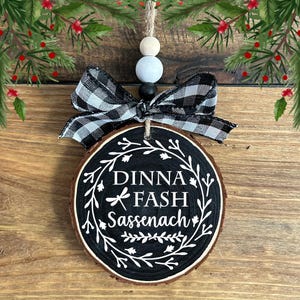 May include: A circular wooden Christmas ornament with the words "DINNA FASH Sassenach" in white script on a black base. It features a black and white checked bow and a hanging string. The ornament is a festive decoration.