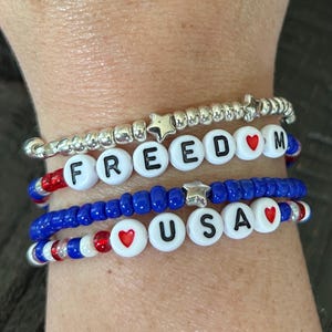 Puede incluir: Un conjunto de pulseras de cuentas que deletrean "FREEDOM" y "USA". Las pulseras presentan cuentas de letras blancas, cuentas de corazón rojas y cuentas azules. Una pulsera es plateada con cuentas en forma de estrella.