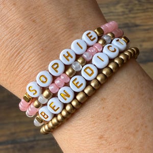 May include: A set of three stacked bracelets featuring round beads in metallic gold and translucent pink. Two bracelets include white circular beads with gold lettering spelling out the names Sophie and Benedict, interspersed with small gold spacers.