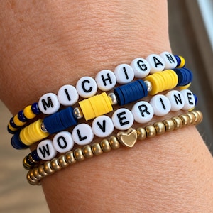 Puede incluir: Una pila de tres pulseras de cuentas. La pulsera superior tiene cuentas de letras blancas que deletrean MICHIGAN con discos azules y amarillos. La pulsera central deletrea WOLVERINE con discos azules y amarillos. La pulsera inferior tiene cuentas doradas metálicas y un dije de corazón.