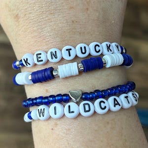 Puede incluir: Tres pulseras de cuentas. La pulsera superior deletrea "KENTUCKY" con cuentas de letras blancas. La pulsera del medio tiene cuentas azules y blancas. La pulsera inferior deletrea "WILDCATS" con cuentas de letras blancas y un dije de corazón plateado.