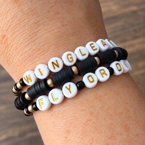 May include: A set of three stretch bracelets featuring white round beads with gold lettering spelling WINGLEA and FLY OR DIE, interspersed with matte black disc beads, small metallic gold spheres, and tiny black round spacer beads.