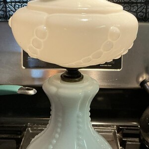 May include: A white milk glass lamp featuring a bulbous base with vertical beaded detailing and a matching wide, rounded shade. The lamp has a metallic brass-toned socket at the top and a circular brass-toned base at the bottom with a power cord.