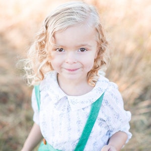 Pode incluir: Uma menina pequena com cabelo loiro encaracolado sorri para a câmera. Ela está vestindo uma blusa branca com gola verde e suspensórios verdes.