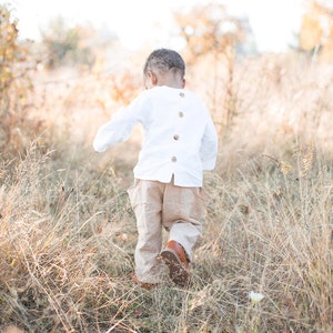 Puede incluir: Un niño pequeño con una camisa blanca abotonada y pantalones beige camina por un campo de hierba alta.
