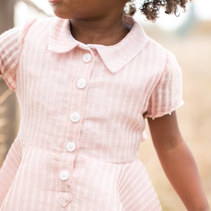 May include: A pink and white striped collared shirt with a button-up front. The shirt has a ruffled hem and short sleeves.