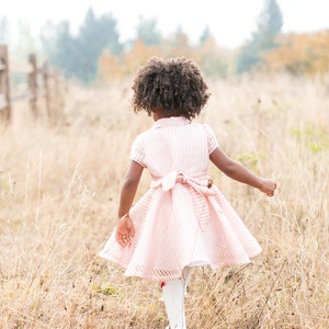 May include: A young girl with curly brown hair wearing a pink and white striped dress walks through a field of tall dry grass.