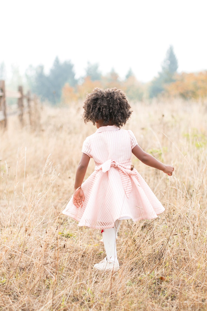 May include: A young girl with curly brown hair wearing a pink and white striped dress walks through a field of tall dry grass.
