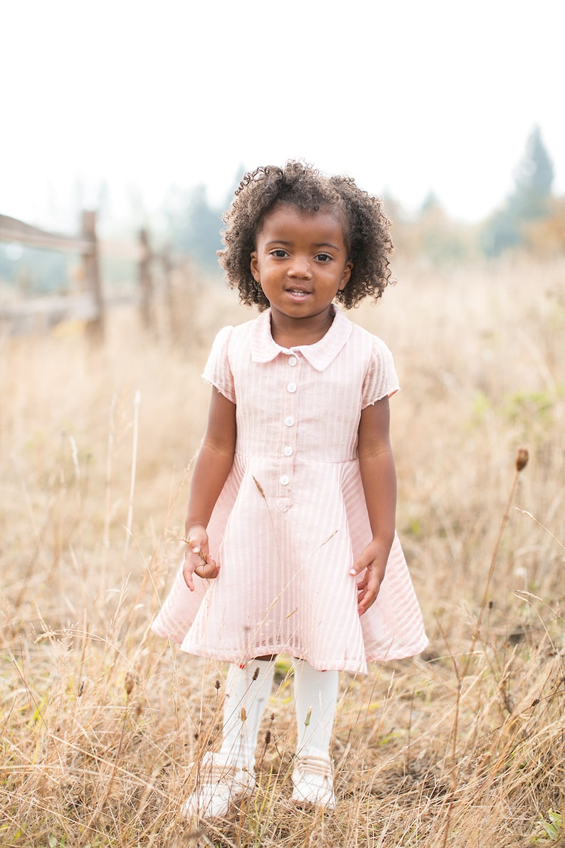 May include: A young girl with curly brown hair wearing a pink and white striped dress with a white collar and buttons. She is standing in a field of long grass.