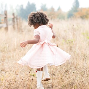 May include: A young girl with curly brown hair wearing a pink and white striped dress and white boots spins in a field of long grass.