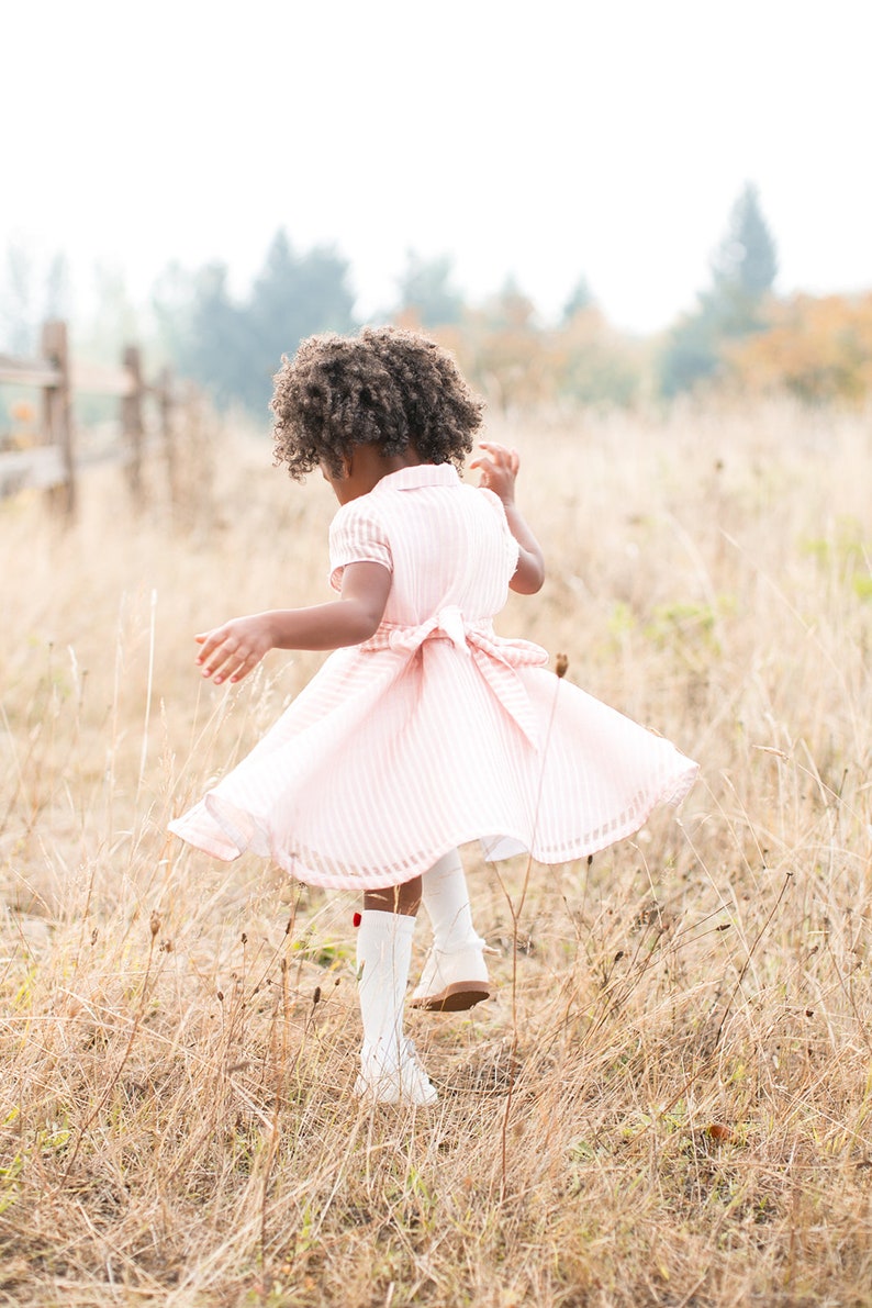May include: A young girl with curly brown hair wearing a pink and white striped dress and white boots spins in a field of long grass.